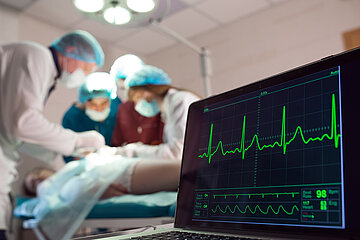 &Auml;rzte operieren einen Patienten, im Vordergrund werden die Vitalwerte auf einem Monitorangezeigt.