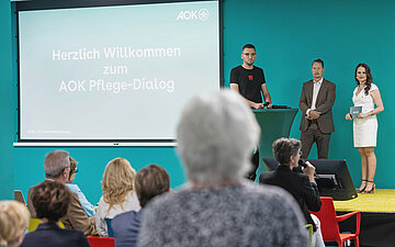 Blick aus dem Publikum auf eine B&uuml;hne beim AOK Pflege-Dialog. Auf der Projektionsfl&auml;che steht &bdquo;Herzlich Willkommen zum AOK Pflege-Dialog&ldquo;; daneben stehen drei Personen vor einer t&uuml;rkisfarbenen Wand.