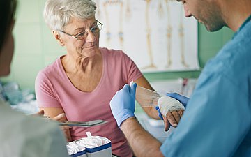Neue Pflegegesetzgebung Foto eines Pflegers, der einer alten Frau in einem Praxisraum einen Verband an der Hand anlegt