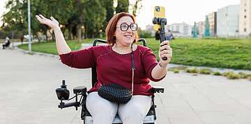 Eine Frau im Rollstuhl h&auml;lt ein Selfie-Stick in ihrer Linken Hand und filmt sich selbst drau&szlig;en auf der Stra&szlig;e. Sie l&auml;chelt, h&auml;lt einen Arm in die Luft und wirkt sehr fr&ouml;hlich.