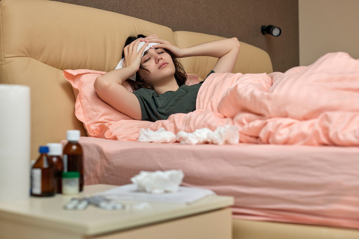 Foto zeigt eine kranke Frau, die im Bett liegt und sich mit beiden Händen den Kopf hält. Auf Ihrem Nachttisch stehen Medikamente und verstreut liegen benutzte Papiertaschentücher.
