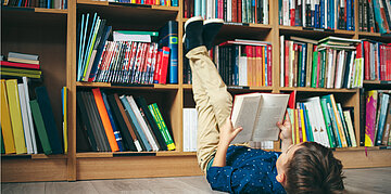 Foto: Ein Kind liegt mit einem aufgeschlagenen Buch in den H&auml;nden auf dem Boden vor einer B&uuml;cherwand.