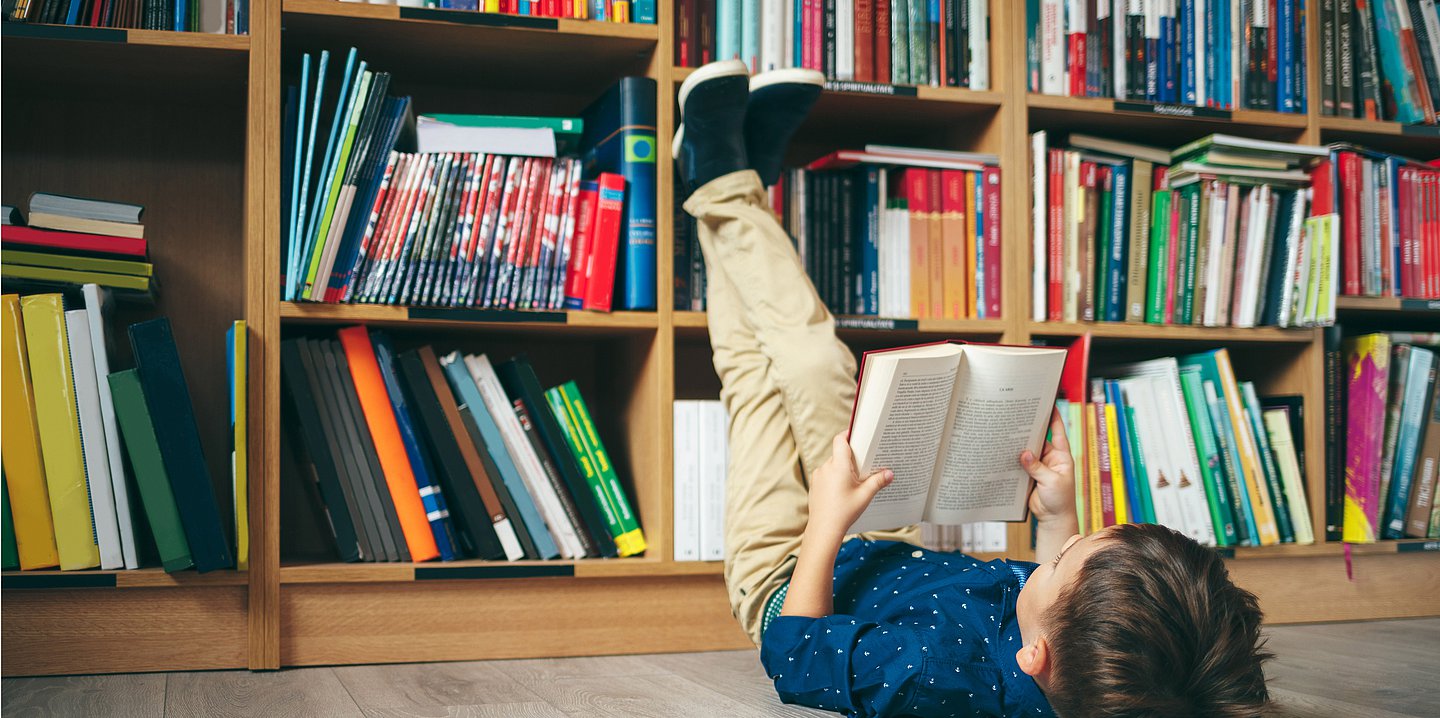 Foto: Ein Kind liegt mit einem aufgeschlagenen Buch in den Händen auf dem Boden vor einer Bücherwand.