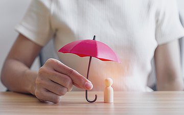 Prävention gegen Gewalt schützt Pflegekräfte Das Bild zeigt eine weibliche Person in einem weißen, kurzärmeligen Kittel. Sie sitzt zum Betrachter gerichtet an einem Tisch. Die rechte Hand ist auf dem Tisch und hält einen kleinen rosafarbenen Schirm über eine kleine Spielfigur.