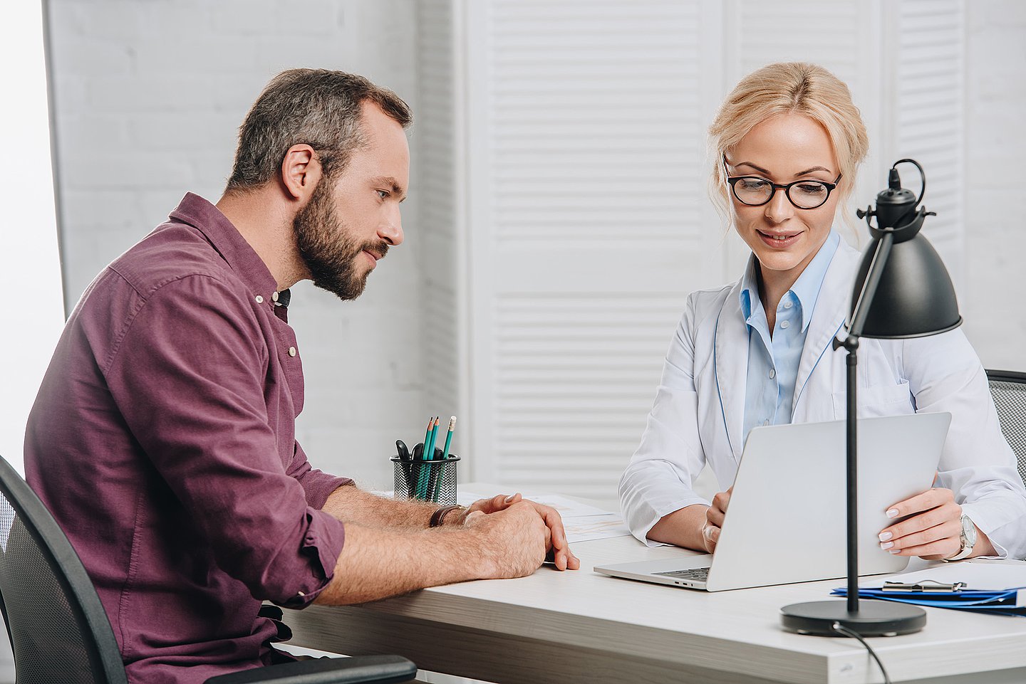 Im Sprechzimmer zeigt eine Ärztin einem Patienten etwas im Laptop, der auf ihrem Schreibtisch steht.