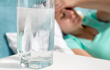 Im Hintergrund liegt eine Frau im Bett und fasst sich an die Stirn, im Vordergrund steht ein Glas Wasser.
