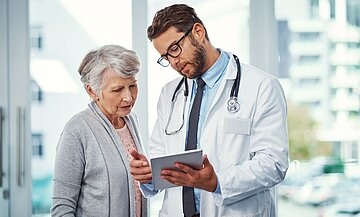Ein Raum mit gro&szlig;en Glasfenstern, davor steht eine &auml;ltere Frau zusammen mit einem j&uuml;ngeren Arzt. Ihm h&auml;ngt ein Stethoskop um den Hals. Beide gucken auf ein Tablet, das der Arzt in der Hans h&auml;lt.
