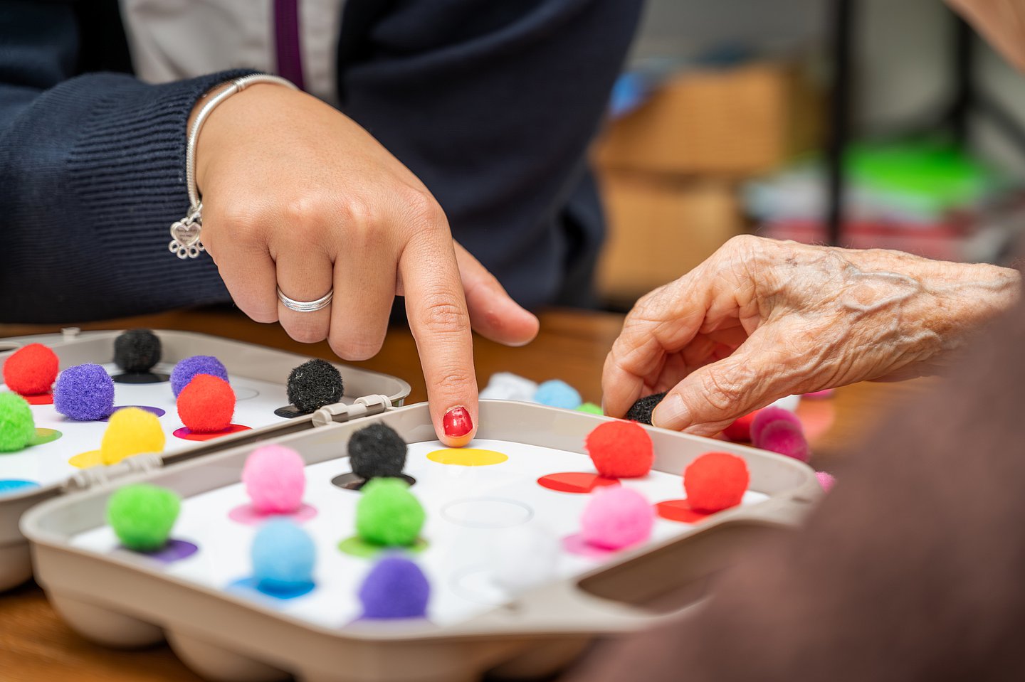 Foto einer pflegebedürftigen älteren Person, die am Tisch sitzt und von einer Therapeutin Unterstützung erhält bei der Zuordnung von kleinen Farbpuscheln auf ein Feld mit farbigen Punkten.