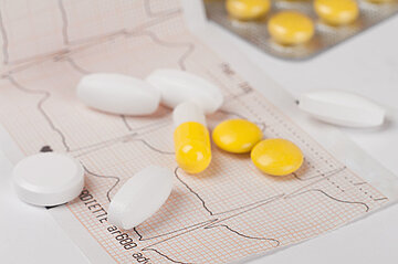 Auf einem Tisch ist ein Teil eines EKG-Protokolls sichtbar, darauf verteilt liegen einige Pillen und Tabletten.