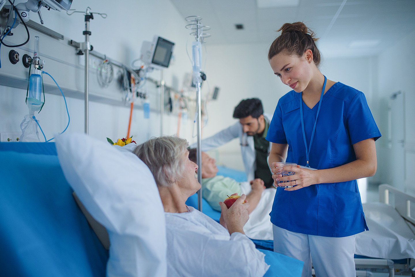 Patienten in einem Behandlungsraum, die von medizinischem Personal betreut werden. Im Vordergrund eine ältere Dame im Bett, die einen Tropf erhält und in den Händen einen Apfel hält; eine Pflegerin bringt ihr ein Glas Wasser.
