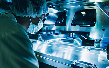 Person in Schutzkleidung und Mundschutz arbeitet an einer Machine in einem pharmazeutischen Produktionslabor.