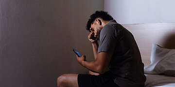Ein Mann sitzt an der Bettkannte, mit dem R&uuml;cken zur Kamera und schaut auf sein Handy. Er hat die rechte Hand an der Stirn und schaut besorgt.