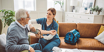 Eine Pflegekraft in medizinischer Kleidung misst bei einem &auml;lteren Mann den Blutdruck in dessen Wohnzimmer. Die beiden sitzen auf einem Sofa.