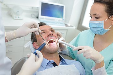 Zahnersatz-Ausgaben in Schleswig-Holstein Ein Patient mit geöffnetem Mund während einer Behandlung beim Zahnarzt auf dem Behandlungsstuhl. Eine zahnärztliche Fachkraft hält auf seiner linken Seite ein Instrument in die Mundhöhle, rechts sind Hände zu sehen, die mit Instrumenten an seinen Zähnen tätig sind.