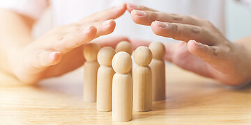 Foto: Sechs Holzspielfiguren stehen auf einem Tisch, dar&uuml;ber h&auml;lt ein Mensch sch&uuml;tzend seine beiden H&auml;nde.