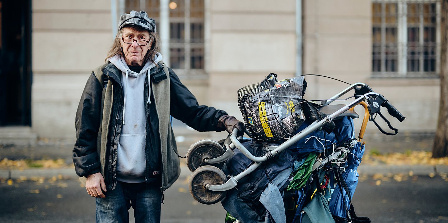 Protagonist Karl steht neben seinem mit Sachen, Regenschirm und Rollator bepackten Wagen