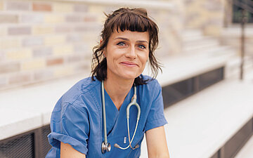 Die Gesundheits- und Krankenpflegekraft, Jenny Kunert sitzt in ihrer Berufskleidung drau&szlig;en auf der Treppe und schaut in die Kamera