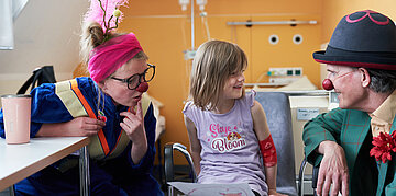 Foto: Ein M&auml;dchen im Krankenzimmer sitzt zwischen zwei Clowns der "Roten Nasen".