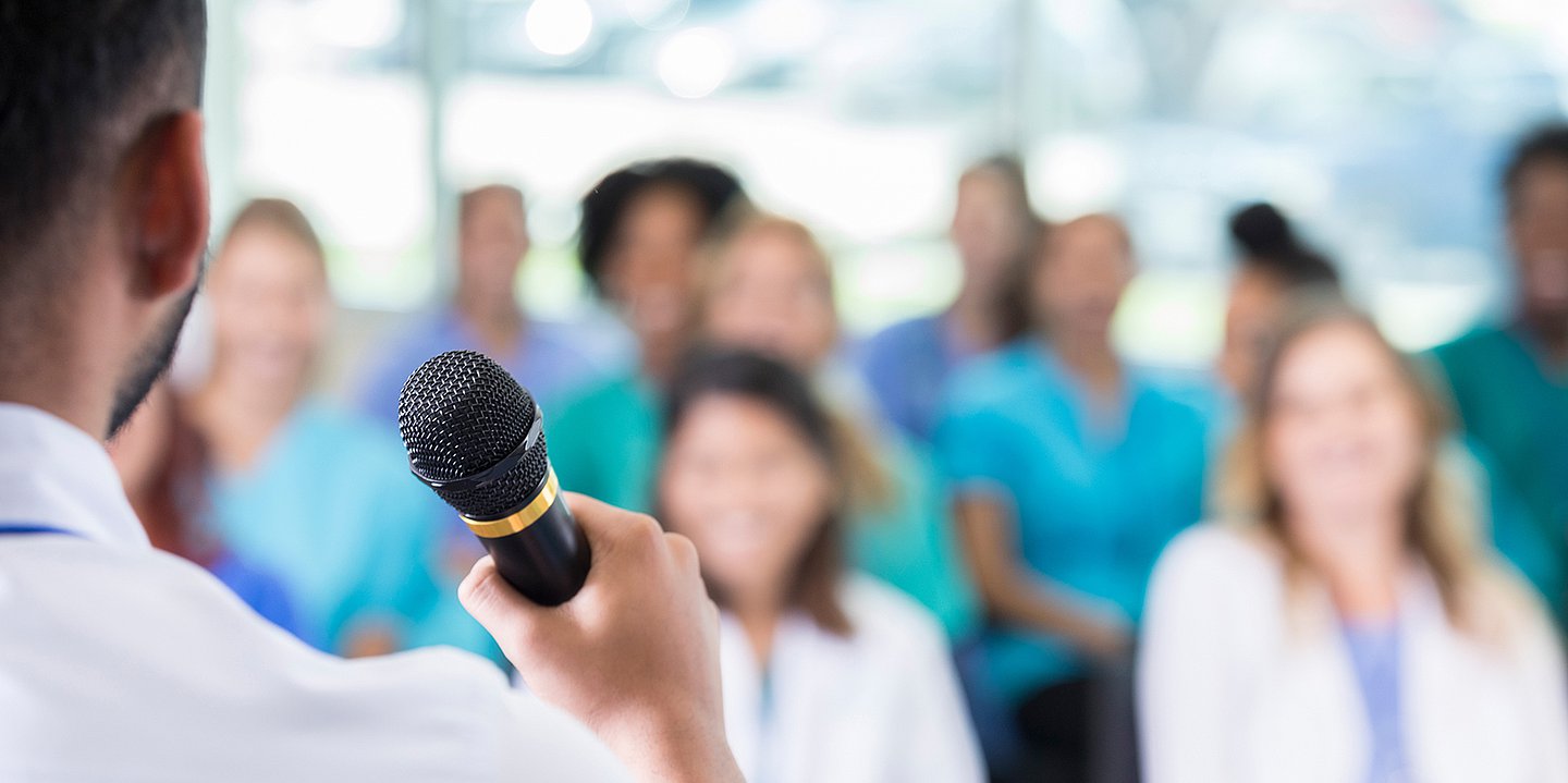 Foto von Medizinerinnen und Medizinern auf einer Konferenz. Im Vordergrund ist von hinten ein Vortragender zu sehen.