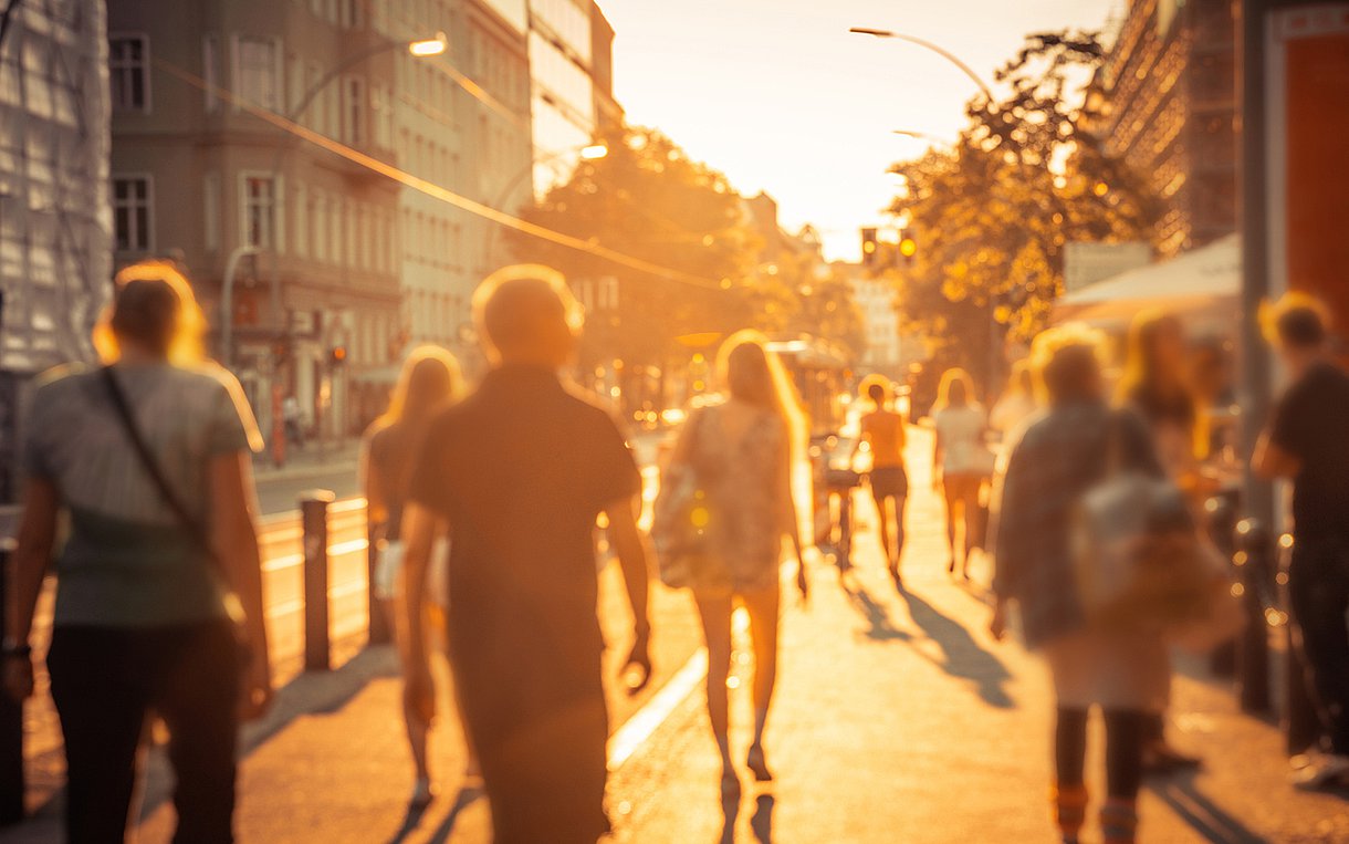 Foto von meheren Fußgängerinnen und Fußgängern, die stark von der Sonne beschienen durch die Stadt laufen. Sie sind nur verschwommen und von hinten zu sehen.