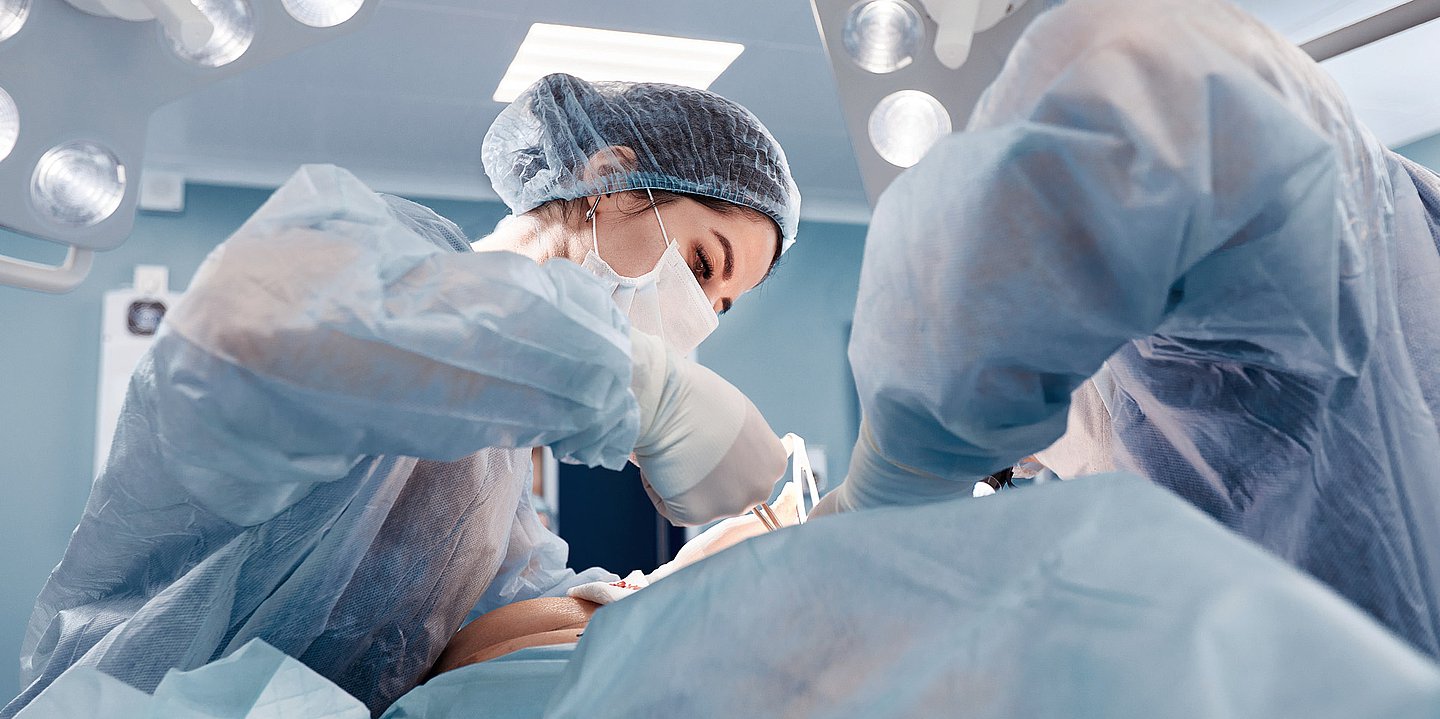 Ärztinbei der OP OP-Szene: Ärztin ist über den Patienten auf dem OP-Tisch gebeugt, recht daneben der Assitent mit dem Rücken zum Betrachter, Perspetive von rechts unten nach links oben