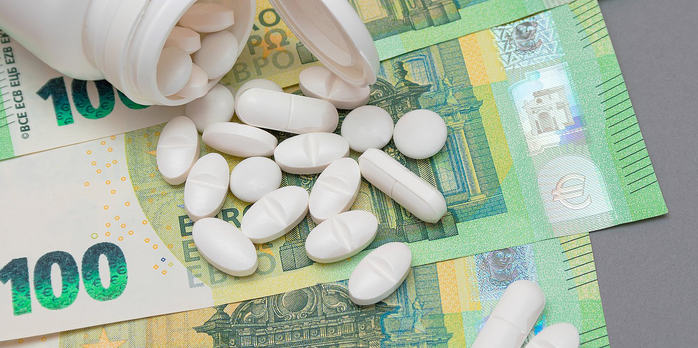 Auf einem Tisch liegen 100-Euro-Scheine. Auf ihnen ist eine Pillenbox umgefallen und ausgeschüttet. Die Pillen liegen verteilt auf den Geldscheinen.