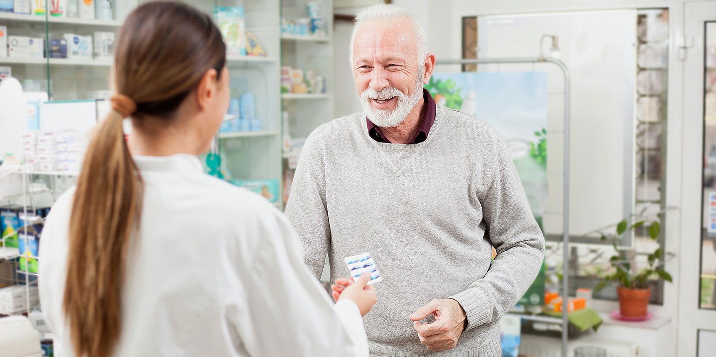 Foto: Ein älterer Mann steht einer Apothekerin gegenüber, die ihm eine Medikamentenschachtel entgegenstreckt.