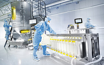 Mehrere Fachkr&auml;fte in Schutzkleidung bedienen Maschinen in einem sterilen Reinraum zur Herstellung pharmazeutischer Produkte.