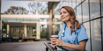 Eine Krankenpflegerin sitzt mit einem to go-Kaffeebecher vor der Einrichtung auf einer Bank und macht Pause.