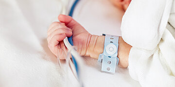 Baby im Krankenhaus: Auf einem wei&szlig;en Laken ist der Arm und die Hand eines Babys zu sehen. Es hat ein blaues Papierarmband um und greift zwei zusammengebundene Schl&auml;uche.