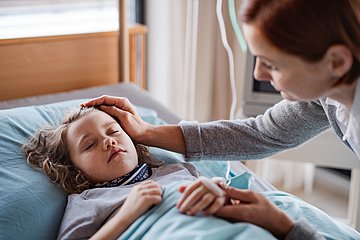 Blinddarm-OPs bei Kindern in Westfalen-Lippe Foto zeigt ein krankes Mädchen, das mit geschlossenen Augen im Bett liegt, die Mutter streicht ihrem Kind sanft über den Kopf.