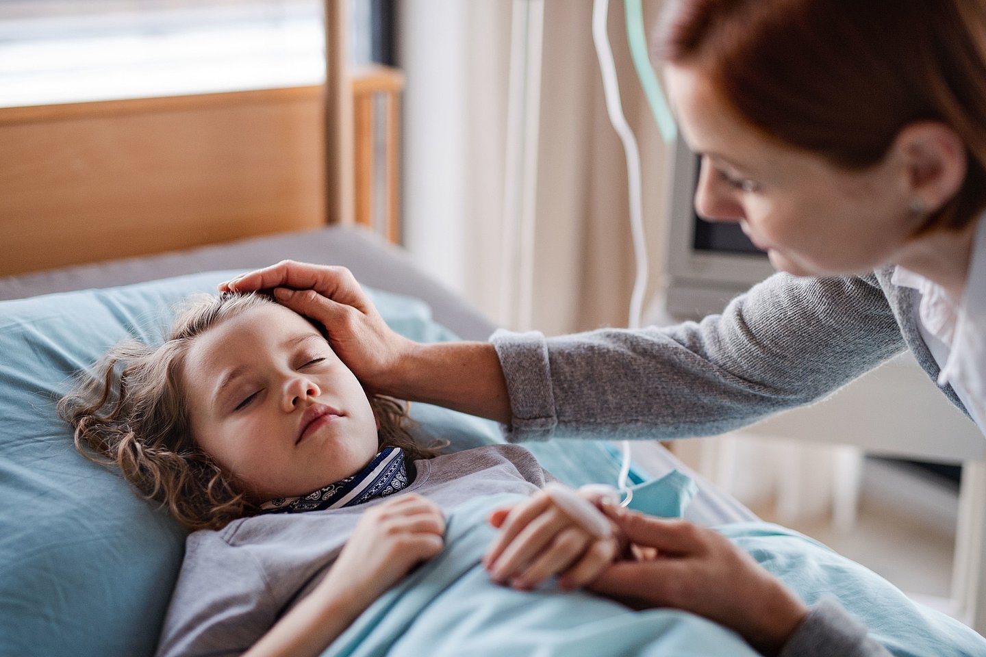 Foto zeigt ein krankes Mädchen, das mit geschlossenen Augen im Bett liegt, die Mutter streicht ihrem Kind sanft über den Kopf.