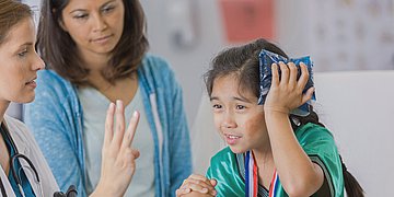 Kind mit Gehirnerschütterung beim Arzt Das Foto zeigt ein dunkelhaariges Mädchen mit geflochtenem Zopf, das sich eine blaue Kaltkompresse an die linke Kopfseite hält. Die Ärztin ihr gegenüber prüft mit dem Anzeigen von drei Fingern die Wahrnehmung des Kindes. Die Mutter steht beobachtend zwischen beiden.