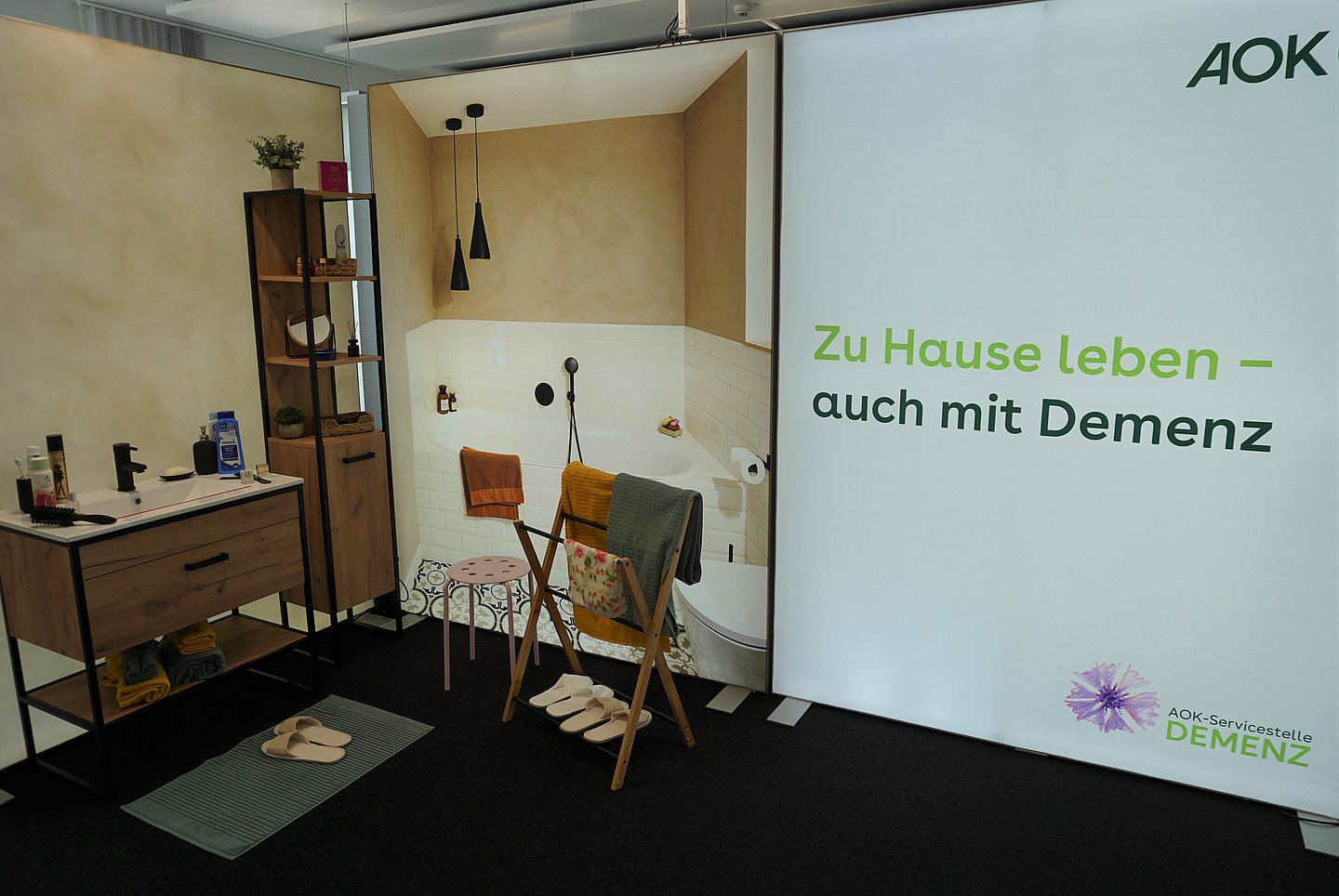 Das Bild zeigt das Badezimmer der Musterwohnung Demenz, das mit einem Waschbecken und einem Foto einer Badewanne gestaltet ist. Zu sehen sind auch ein Handtuchhalter mit Handtüchern sowie ein regal mit verschiedenen Gegenständen.