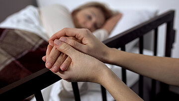 Eine am Bett sitzende Frau h&auml;lt die Hand einer kranken Frau.