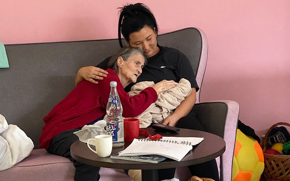 Foto: Zwei Personen, eine davon älter, sitzen auf einem Sofa. Die ältere Person lehnt sich an die jüngere, die sie in den Arm nimmt.