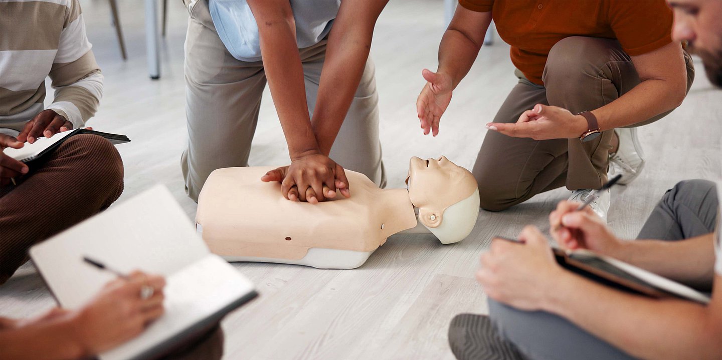 Um eine Übungspuppe sitzen verschiedene Menschen im Kreis. Eine Person übt die Herzdruckmassage.