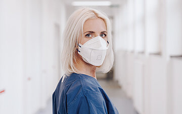 Das Bild zeigt die Gesundheits- und Krankenpflegerin Vanessa Schulte im Klinikflur, zur&uuml;ckschauend in die Kamera mit Maske
