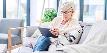 Foto: Eine Frau mittleren Alters sitzt auf einem hellen Sofa und h&auml;lt ein Tablet in die Hand, in das sie schaut.