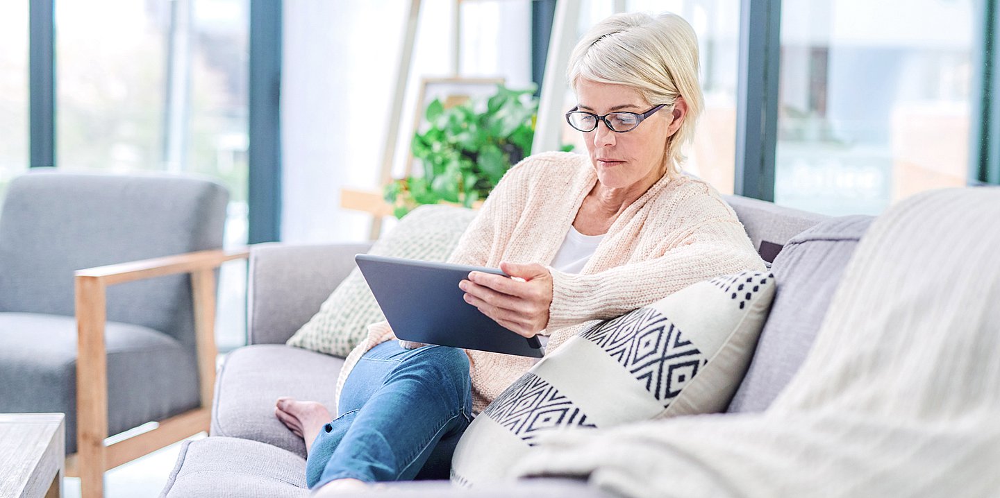 Foto: Eine Frau mittleren Alters sitzt auf einem hellen Sofa und hält ein Tablet in die Hand, in das sie schaut.