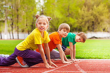 Das Foto zeigt ein (Schul-)Sportgel&auml;nde; im Vordergrund sind eine Sch&uuml;lerin und zwei Sch&uuml;ler zu sehen, die auf einer Tartanbahn die Startposition zum Loslaufen eingenommen haben und l&auml;chelnd in die Kamera schauen.