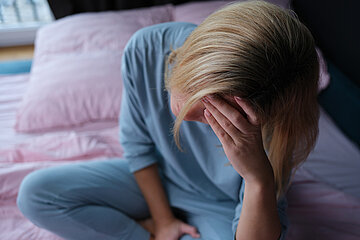 Ein Frau in einem blauen Schlafanzug sitzt mit gekreuzten Beinen auf ihrem Bett, die linke Hand an den Kopf gest&uuml;tzt, den Blick nach unten gerichtet.