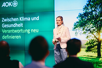 Susann Hinz, Personalmanagerin der AOK Hessen, steht vor einer Videowand, die einen Baum in der Landschaft abbildet und h&auml;lt einen Vortrag