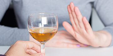 Foto einer Frau, der ein Glas Wein zugeschoben wird und die dankend mit einer abwehrenden Handgeste ablehnt