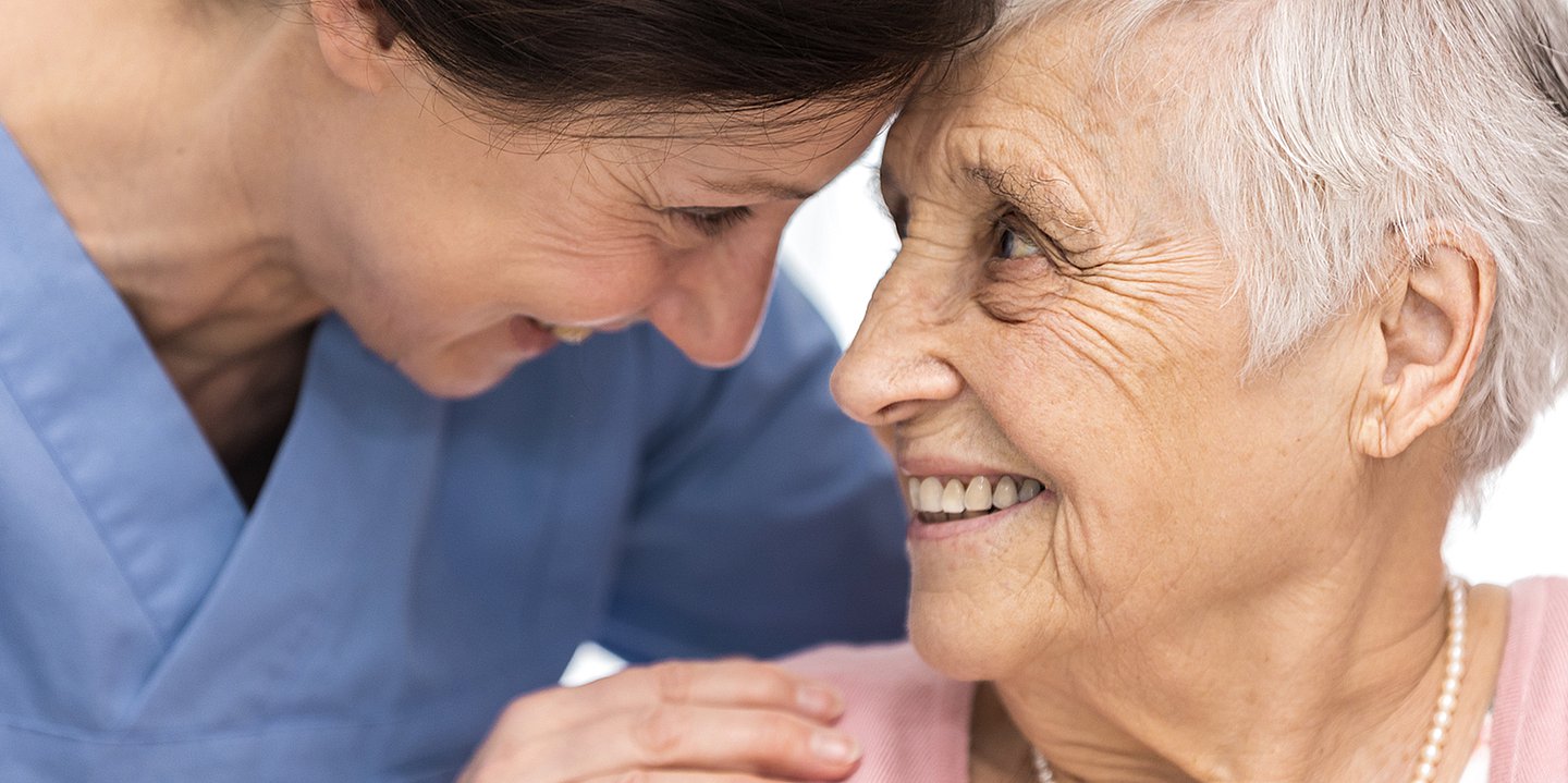 Foto: Eine jüngere Frau in blauer Pflegetracht drückt eine ältere Frau an der Schulter und lächelt sie freundlich an.