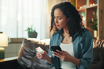 Eine Frau sitzt auf dem Sofa im Wohnzimmer, sie schaut auf ein Medikament, dass sie in linken Hand h&auml;lt. In der rechten Hand hat sie ihr Handy, mit dem sie die Informationen abgleicht.