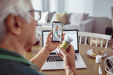 Foto: Ein Senior im Video-Chat mit einem Arzt: In der linken Hand h&auml;lt er ein Smartphone, in der rechten Hand ein Medikament. 