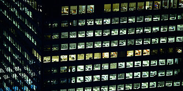 Foto: N&auml;chtliche Ansicht eines B&uuml;rogeb&auml;udes mit zahlreichen beleuchteten Fenstern und sichtbaren Arbeitspl&auml;tzen.