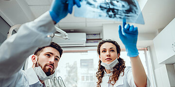 Foto: Zwei junge Mediziner in wei&szlig;er Tracht halten ein R&ouml;ntgenbild hoch &ndash; die Frau zeigt darauf.