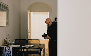 Armin Nassehi steht neben einem Besprechungstisch und blickt auf einen Gegenstand in seinen H&auml;nden. Auf dem Tisch liegen Unterlagen, daneben stehen Getr&auml;nkeflaschen und Gl&auml;ser.